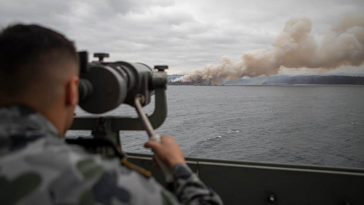 Photo of Seaman Boatswains Mate Malik El-Leissy watches a burning fire from HMAS Adelaide as the ship arrives at Eden to assist with wildfires